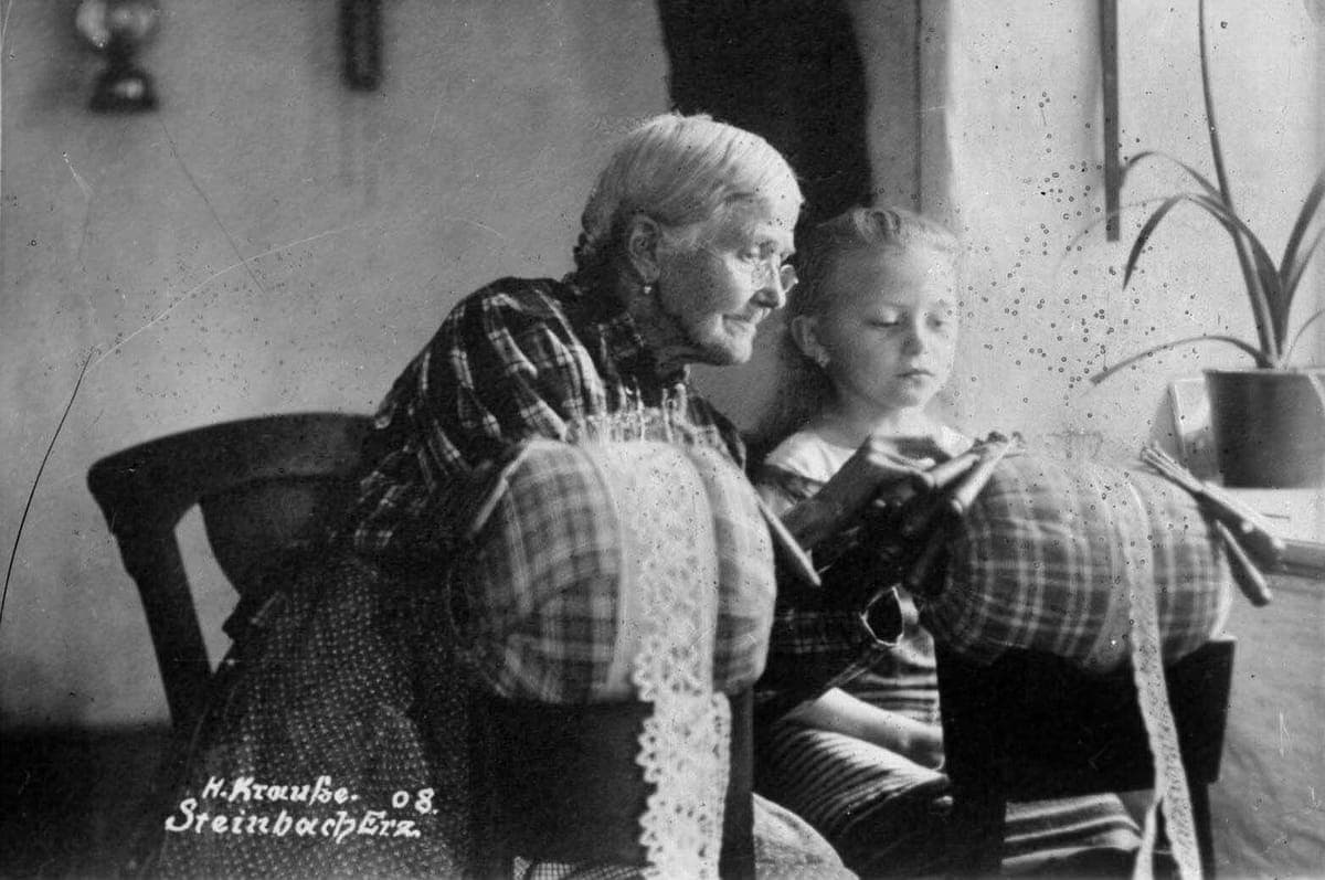 Großmutter mit Brille zeigt ihrer Enkelin das Klöppeln, Steinbach 1908: links das Original in Schwarzweiß mit vielen Stockflecken, rechts mit KI restauriert und koloriert.